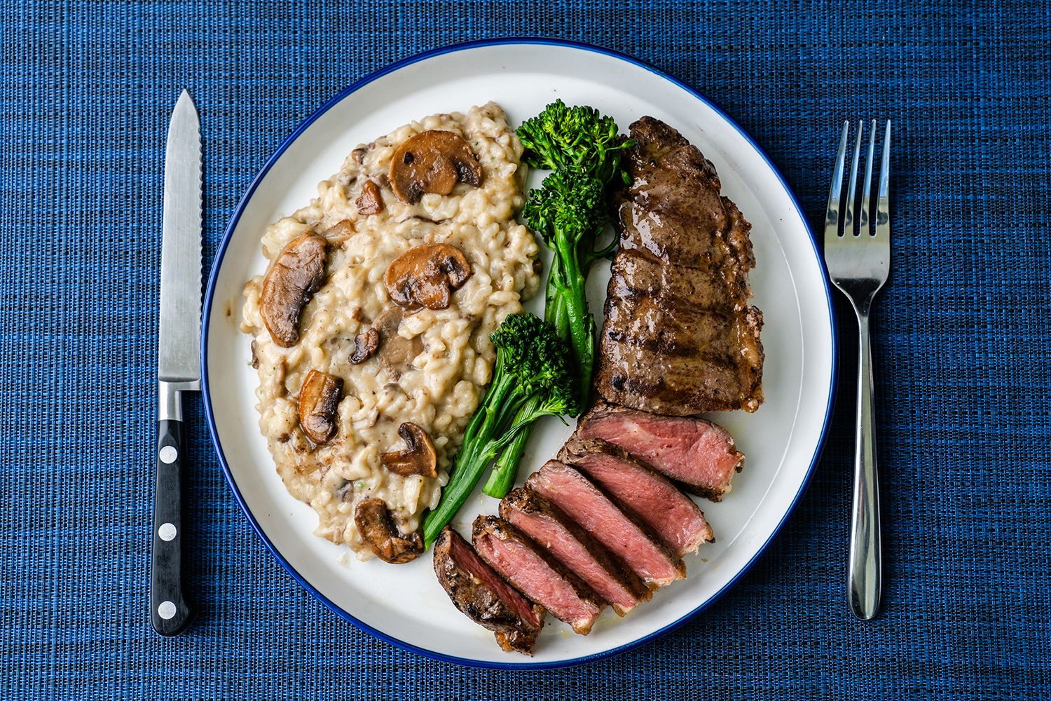 Professionally lit dish of mushroom risotto, broccolini and steak on a white dish with blue background.