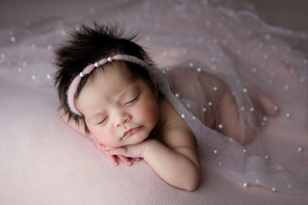 Sleeping newborn with dark hair wearing soft pink headband and tulle wrap.