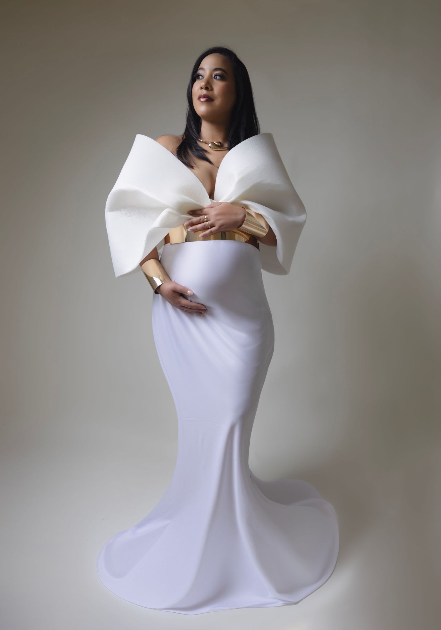 Elegant maternity portrait in a form-fitting white gown with dramatic ruffled shoulder detail against neutral backdrop.