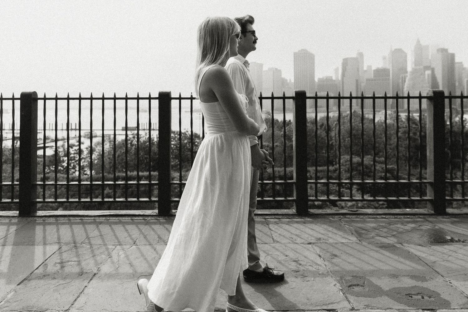 Black and white photo of a enaged couple standing by a railing with Manhattan skyline backdrop. Brooklyn, NY. Miranda Photo & Co