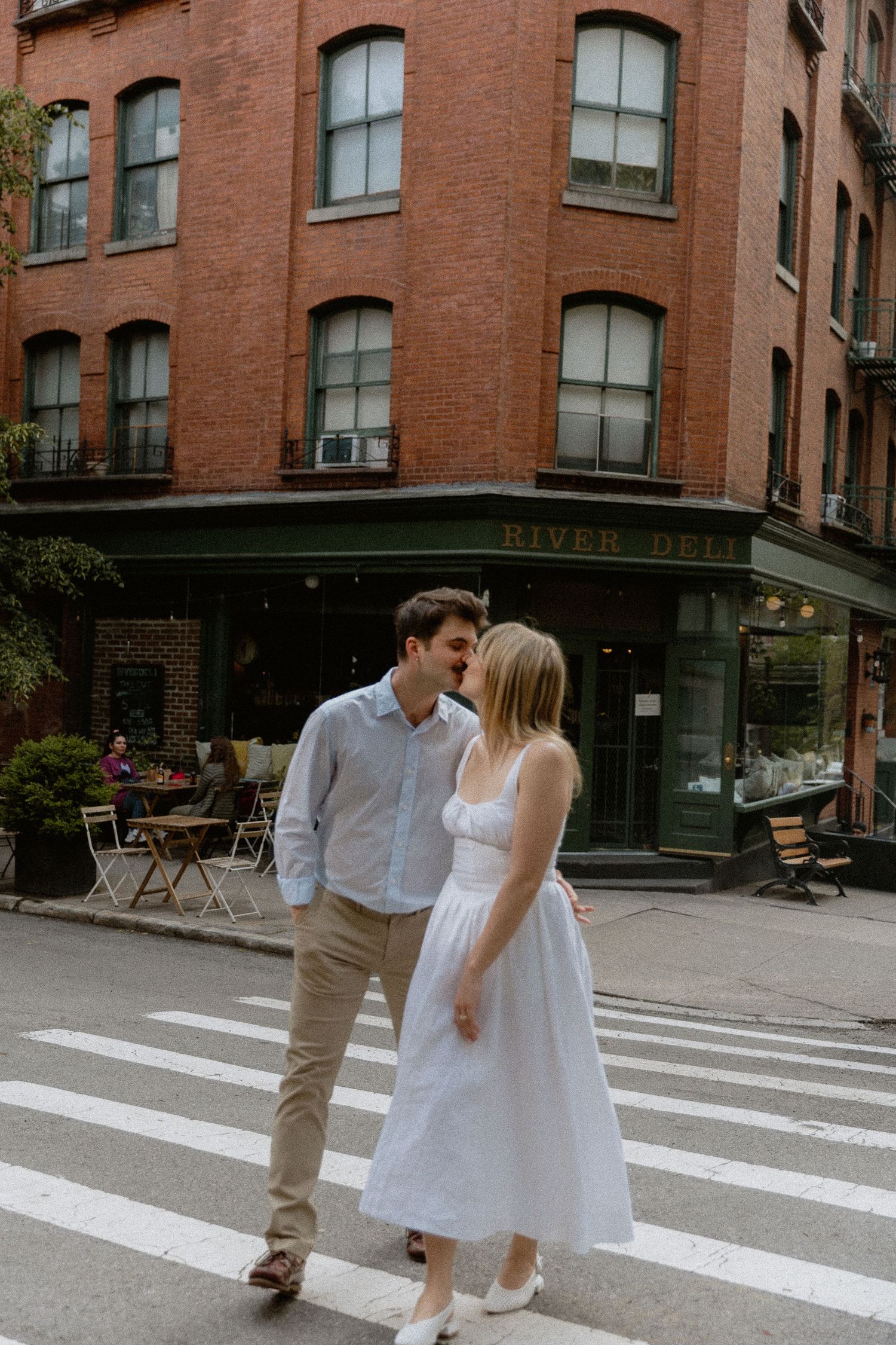 Engaged Couple sharing a romantic kiss while crossing the streen in front of a brick building. Brooklyn, New York. Miranda Photo & Co
