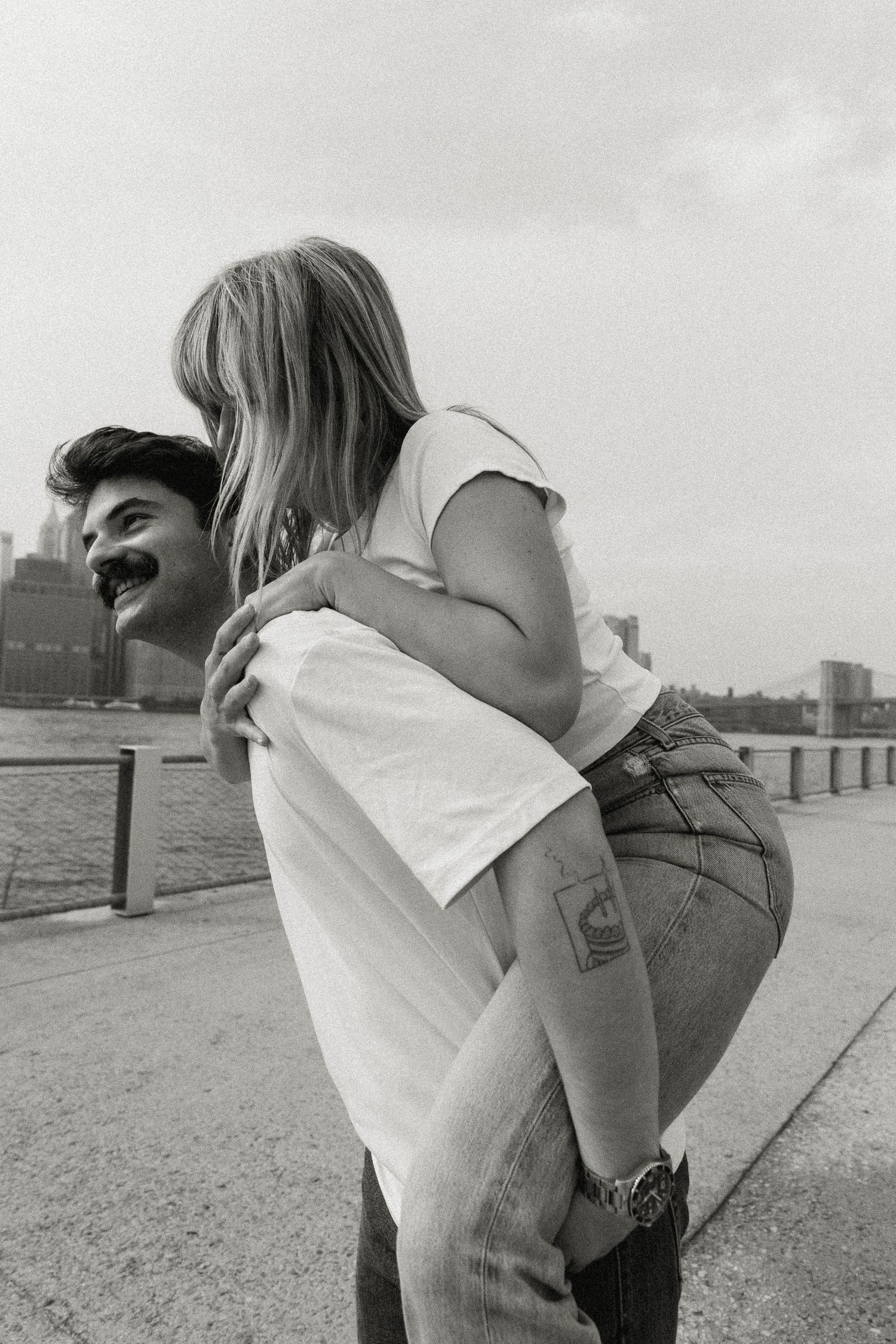 Black and white photo of a man giving a woman a piggyback ride along a waterfront promenade. Brooklyn, New York Miranda Photo & Co
