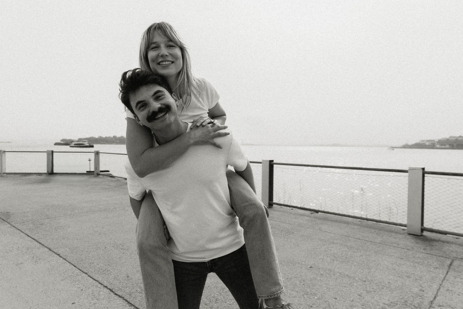 Black and white photo of a joyful engaged couple, man giving woman a piggyback ride by the waterfront. Brooklyn New York Miranda Photo & Co