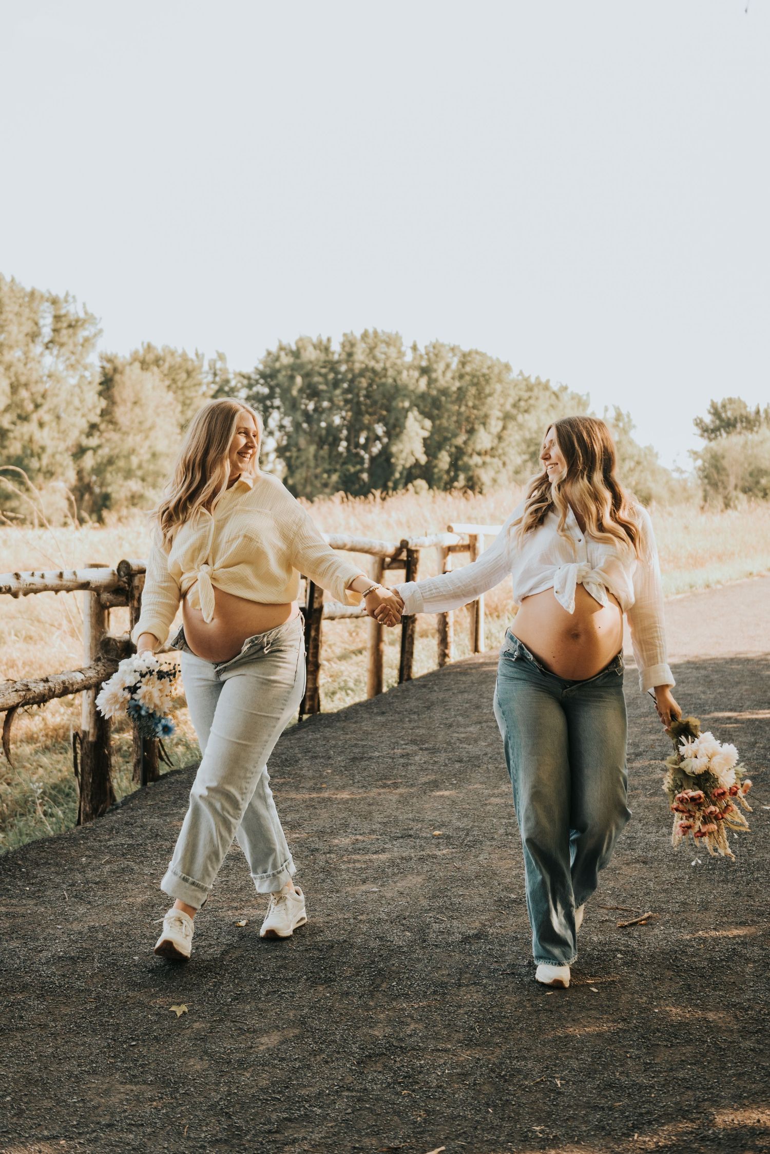 Two women in casual outfits dance together in a field during a golden hour photoshoot.