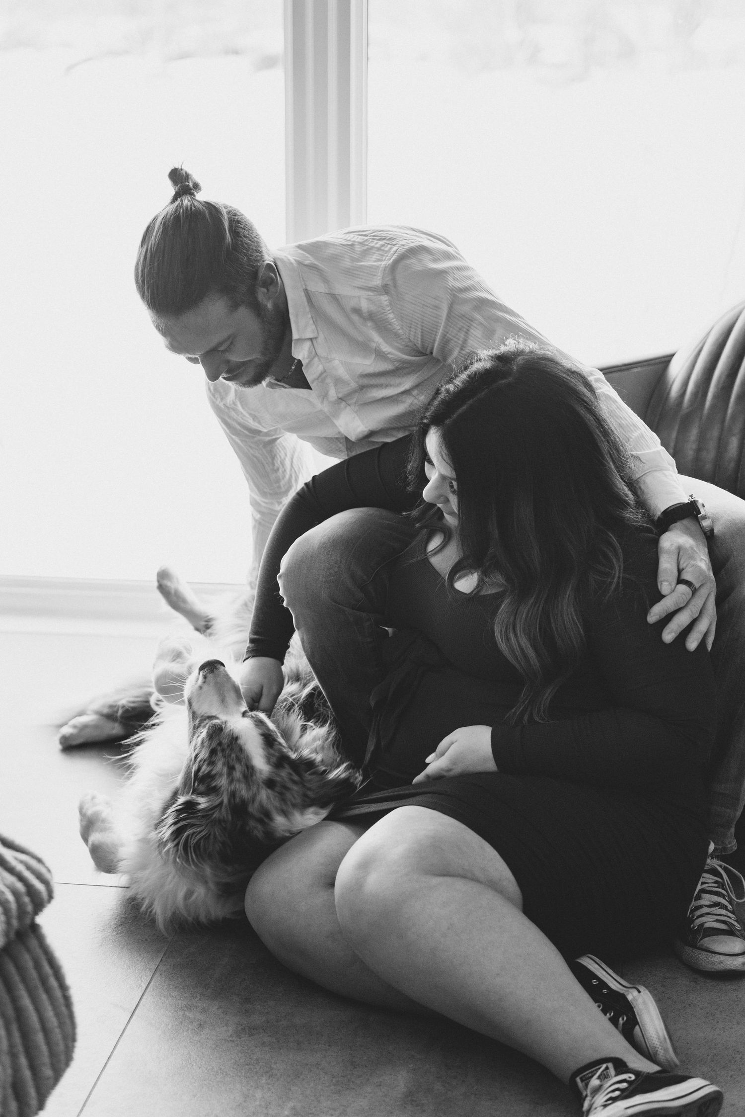 A couple sits on the floor playing with their fluffy dog in a bright room by a window.