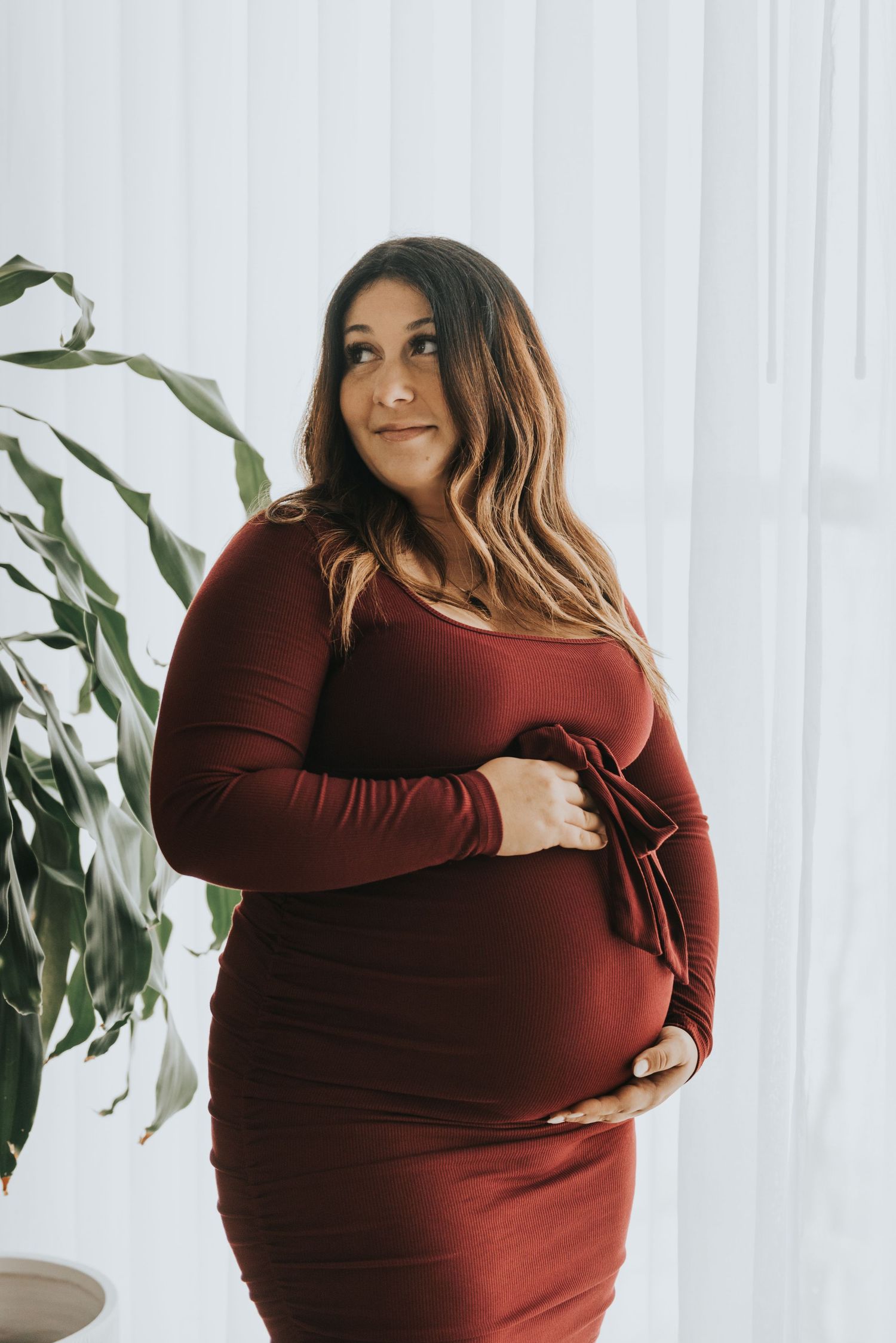 Expectant mother in burgundy form-fitting dress poses by window with indoor plant in natural lighting.
