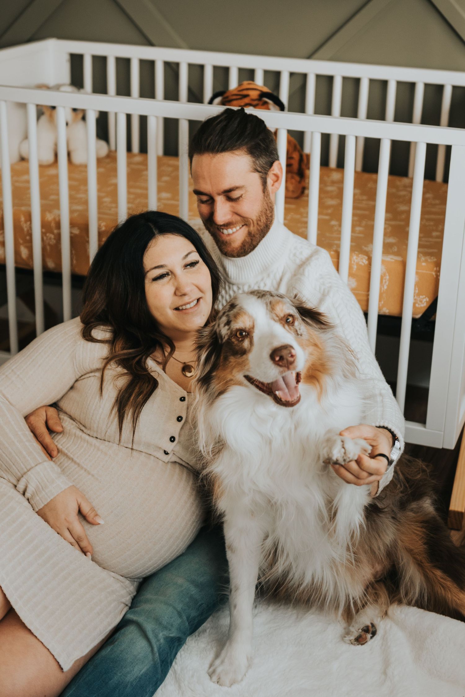A couple relaxes on a couch with their fluffy Australian Shepherd dog in a warm, cozy home setting.