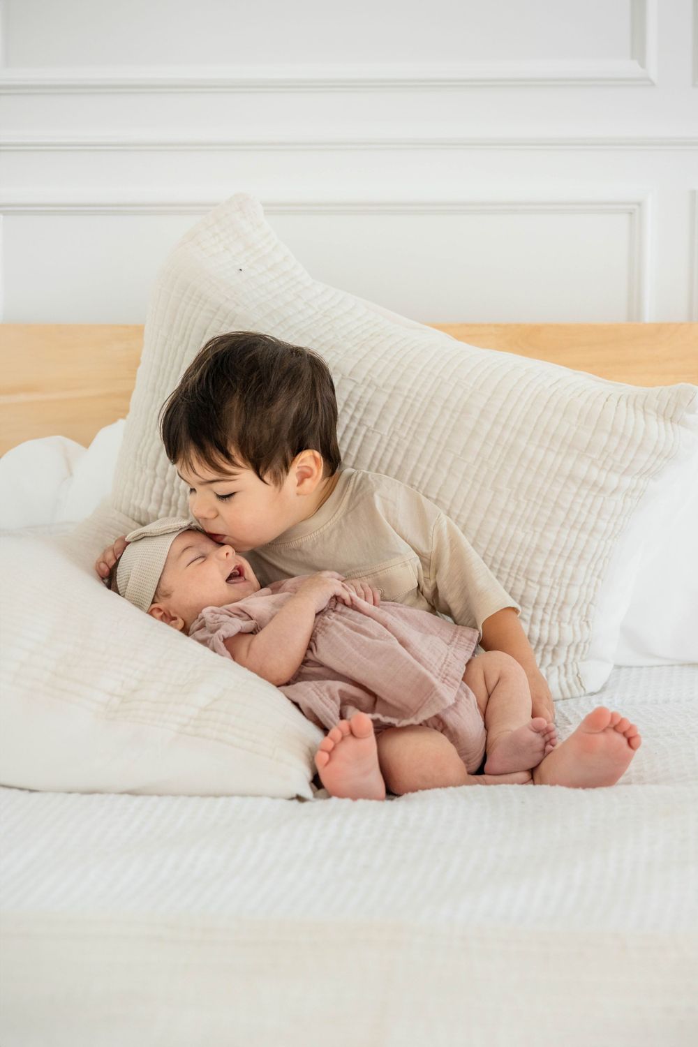 Toddler boy holds and kisses his new baby sister on a bed.