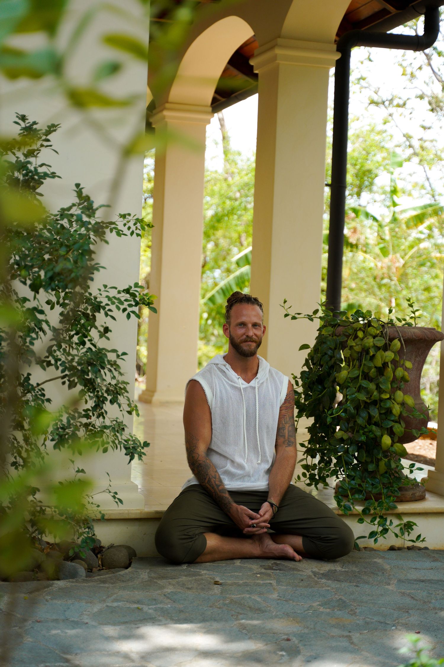 Person meditating in cross-legged pose under arched porch with garden foliage.