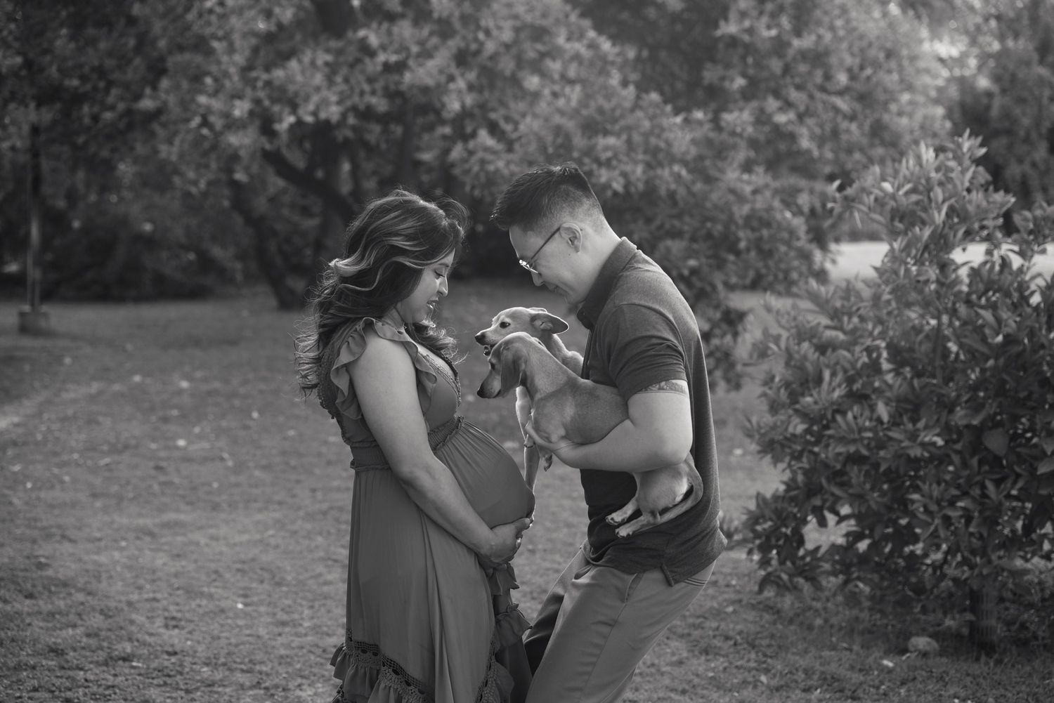 Black and white photo of a pregnant couple with a dog in a lush park setting.