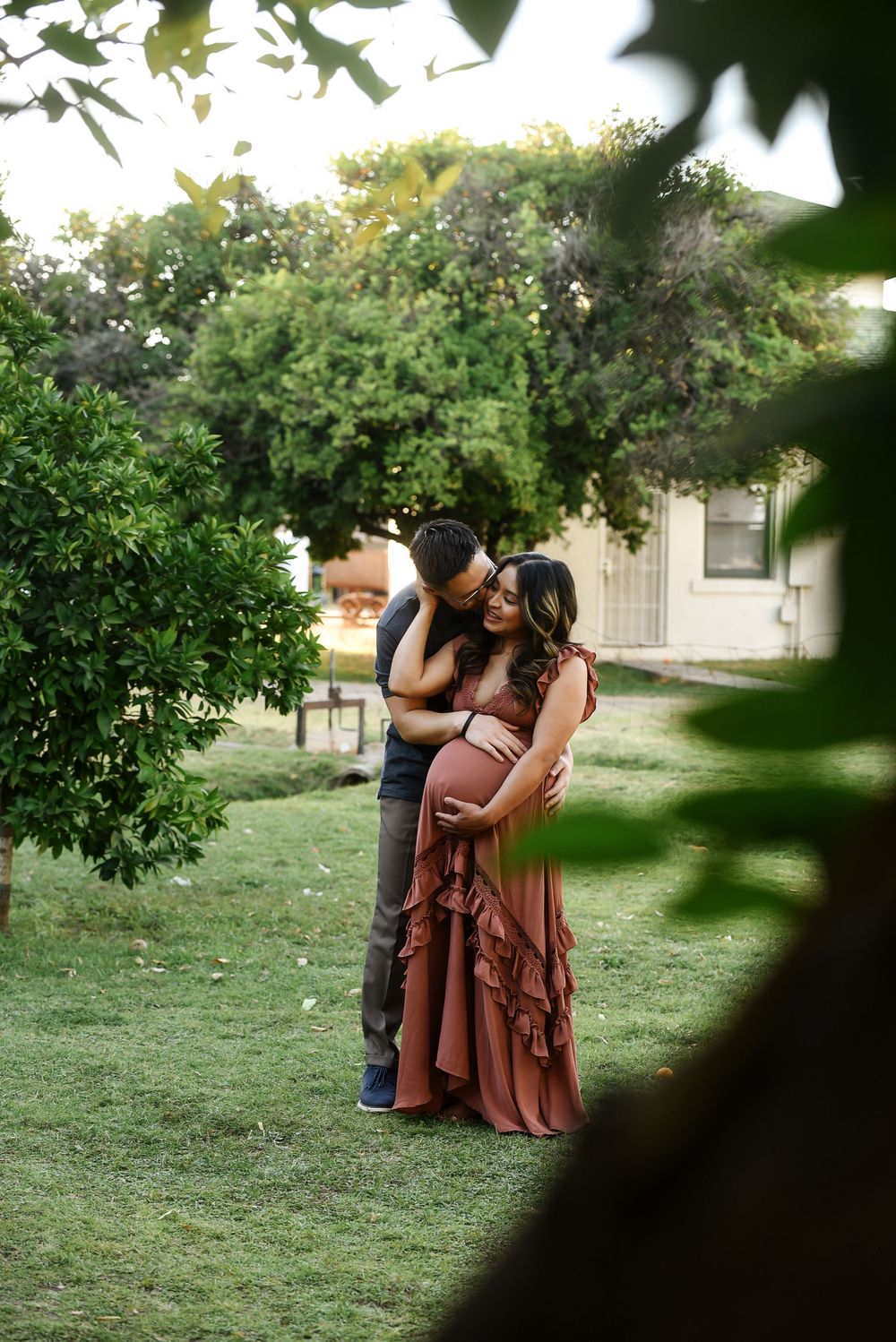 Couple shares tender maternity moment outdoors, man kissing woman's head while cradling her pregnant belly in lush garden.