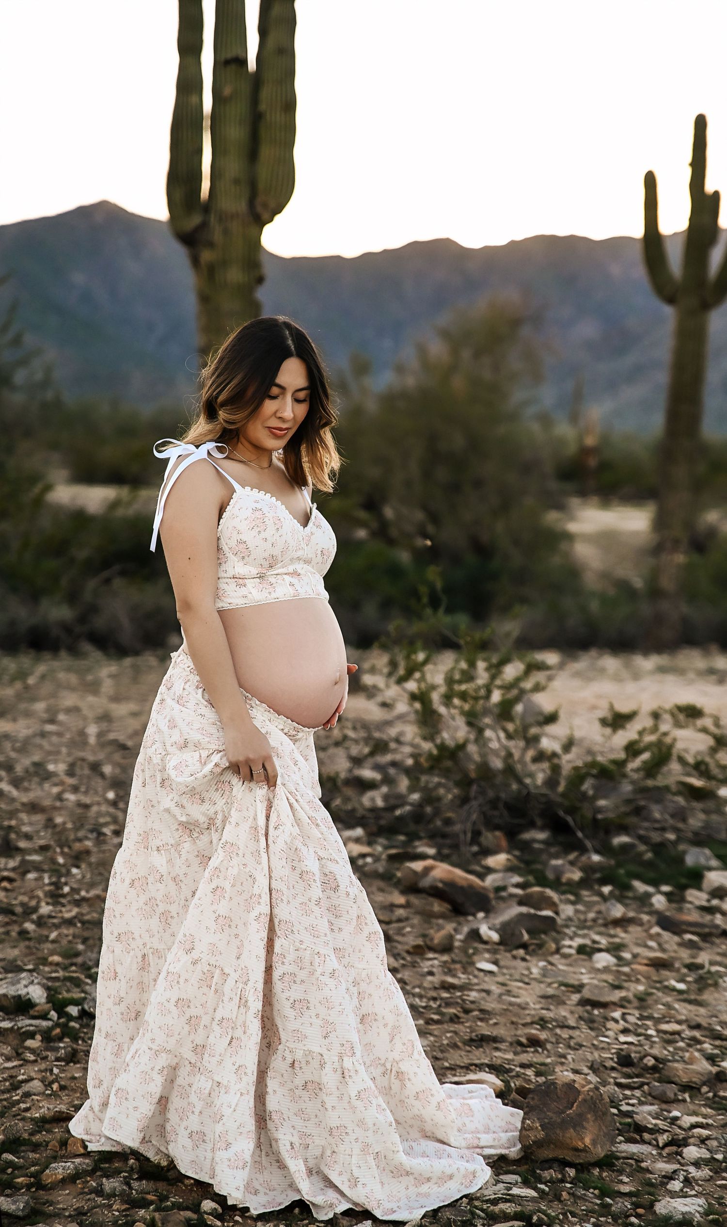 Pregnant woman in floral maternity gown poses in desert landscape with saguaro cacti and mountains at golden hour.