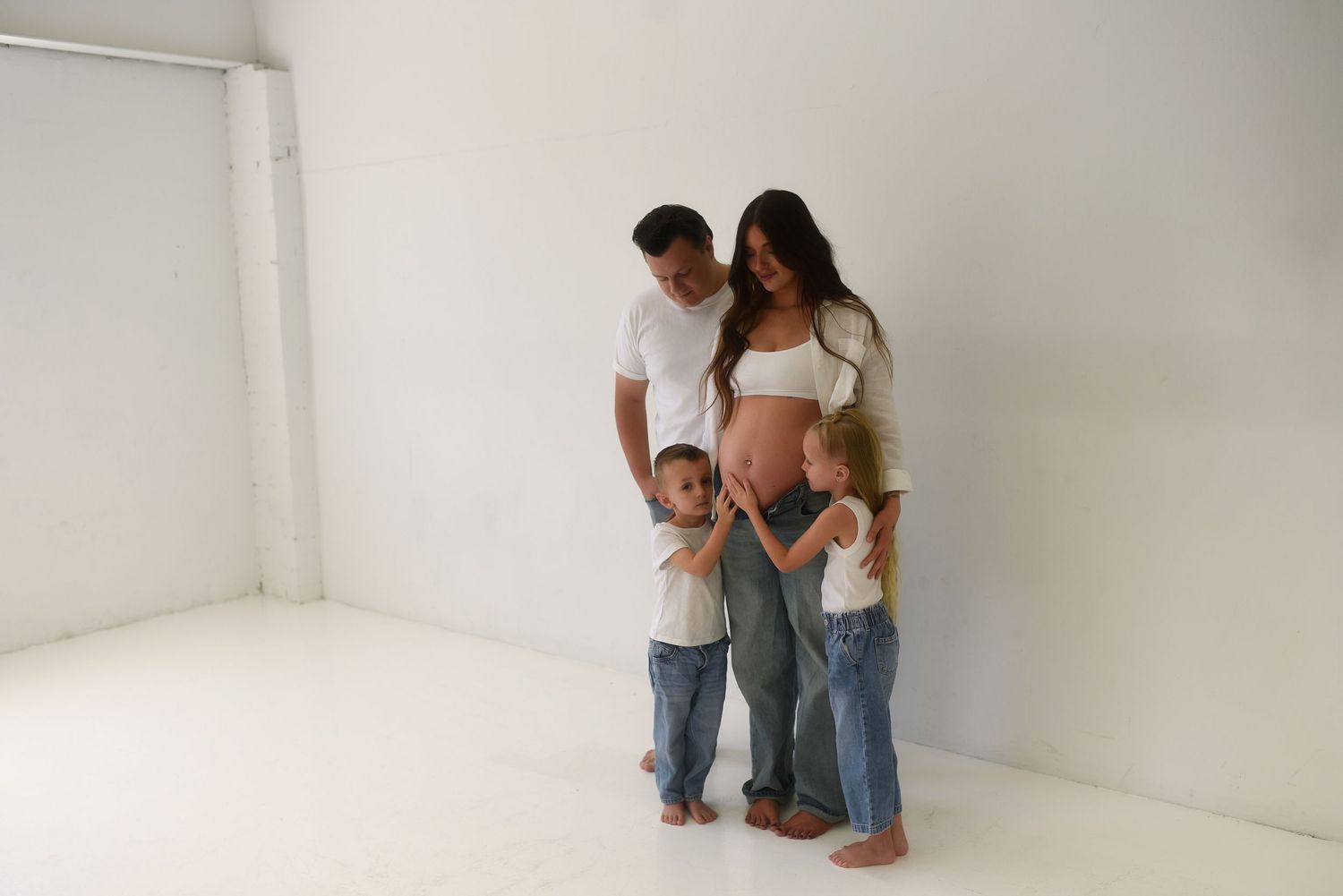 Pregnant woman with partner and two young children posing together in a bright white studio for a maternity photo.