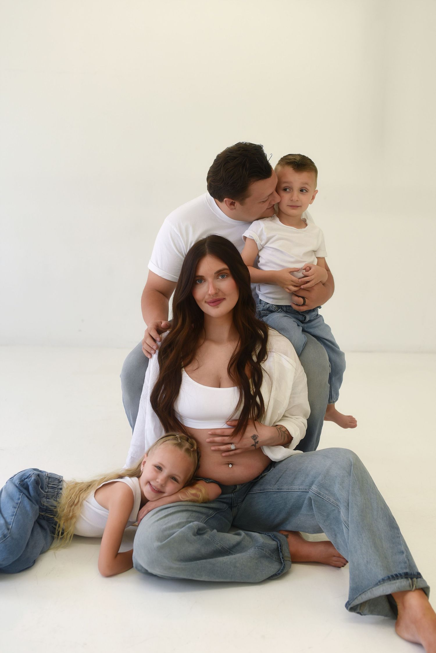 Pregnant woman sits on white floor with long dark hair, husband kisses toddler son, young daughter leans on her lap.