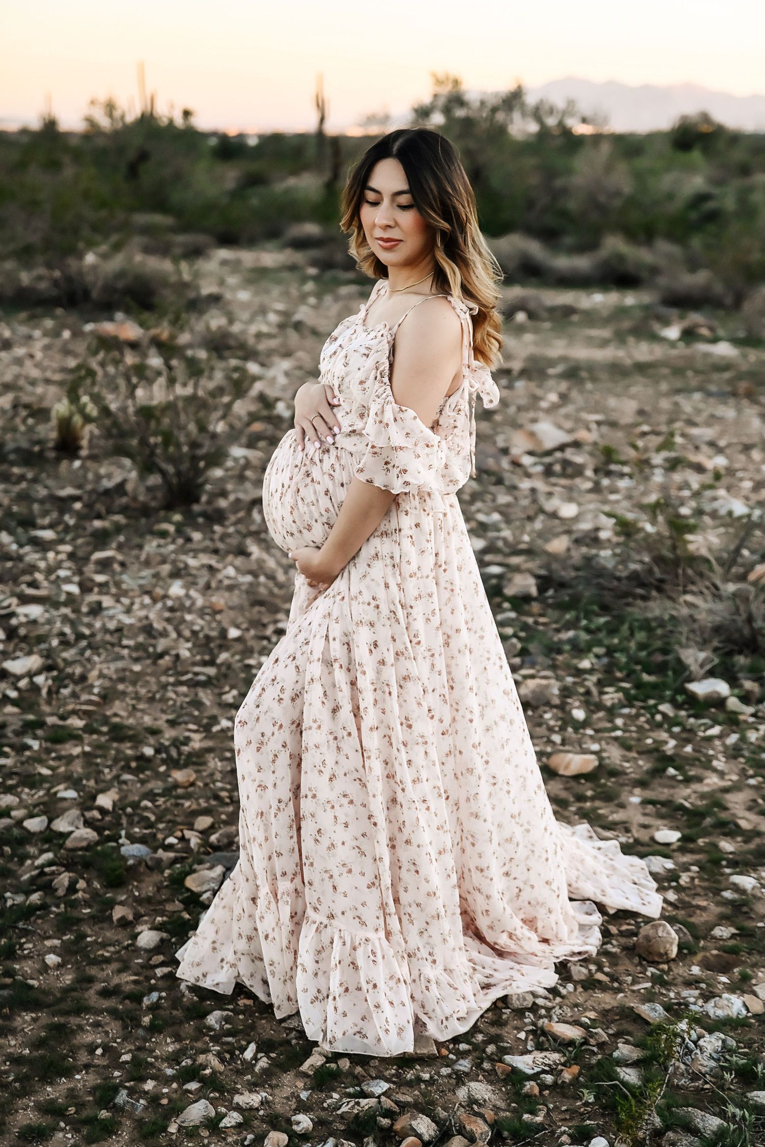 Pregnant woman in floral maxi dress posing outdoors in desert landscape at sunset.