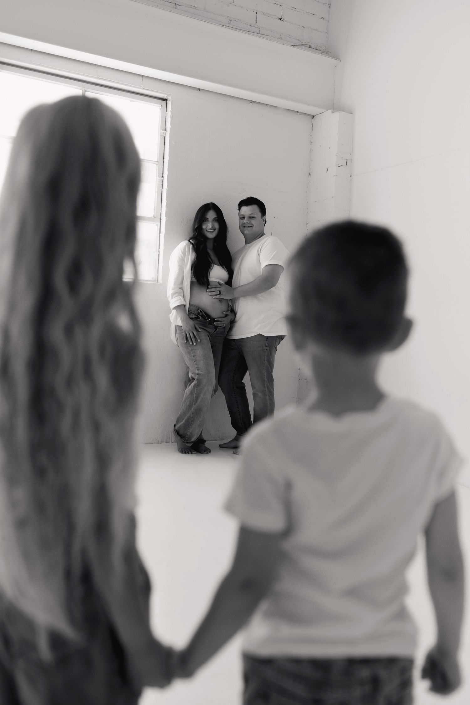 Black and white photo of pregnant couple posing while two children hold hands in foreground.
