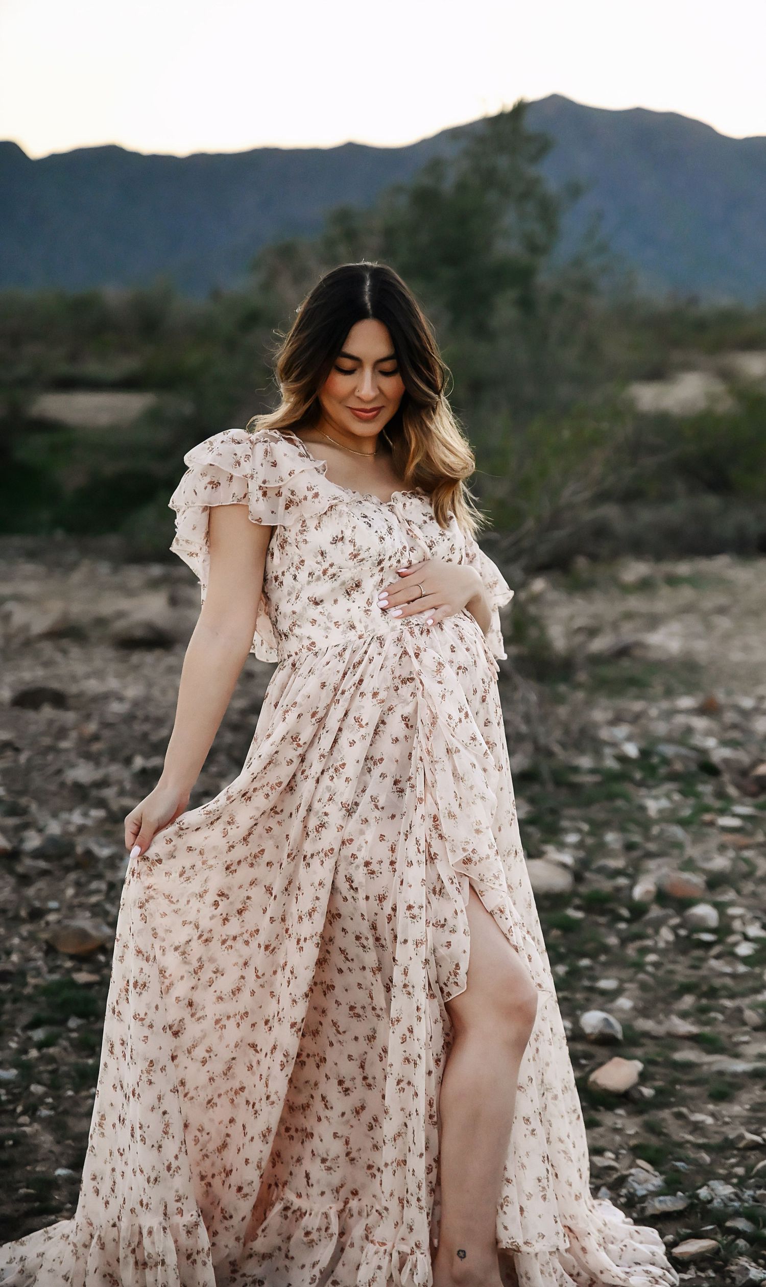 Pregnant woman in floral maxi dress posing outdoors with mountain backdrop during golden hour.