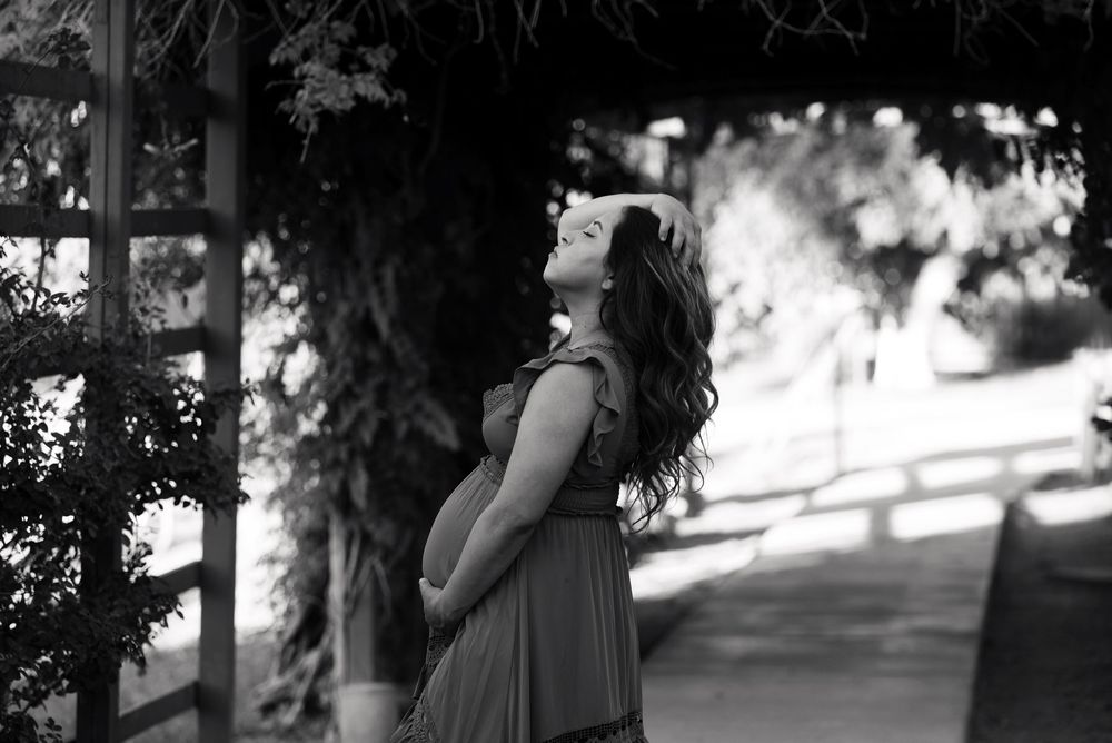Black and white photo of a pregnant woman in a flowing dress, cradling her belly under a vine-covered pergola.