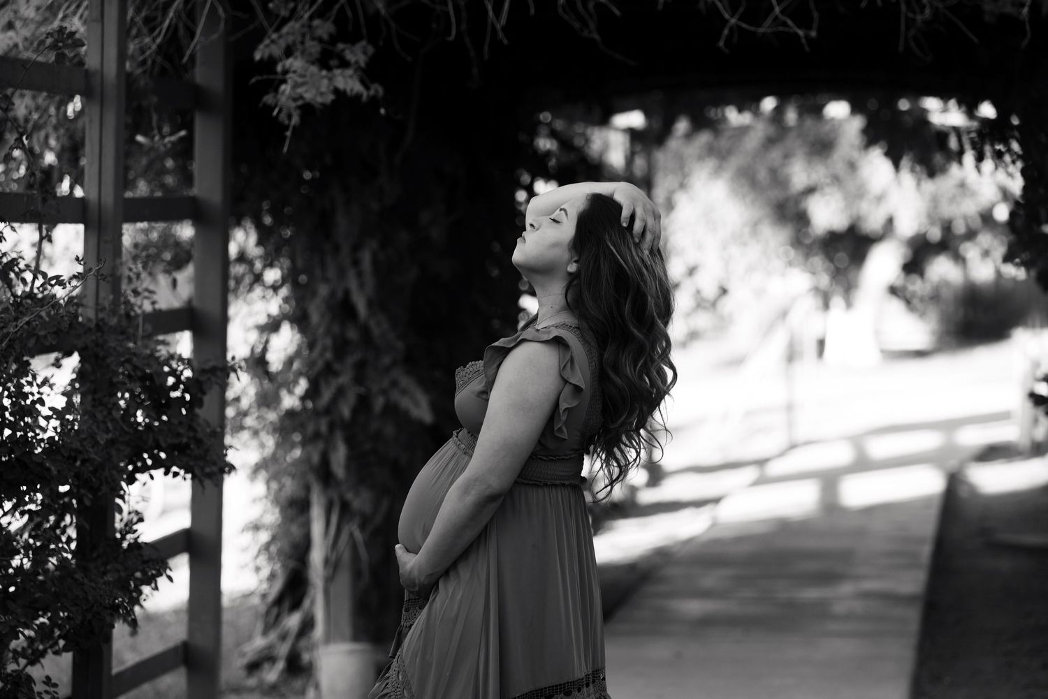 Black and white photo of a pregnant woman in a flowing dress, cradling her belly under a vine-covered pergola.