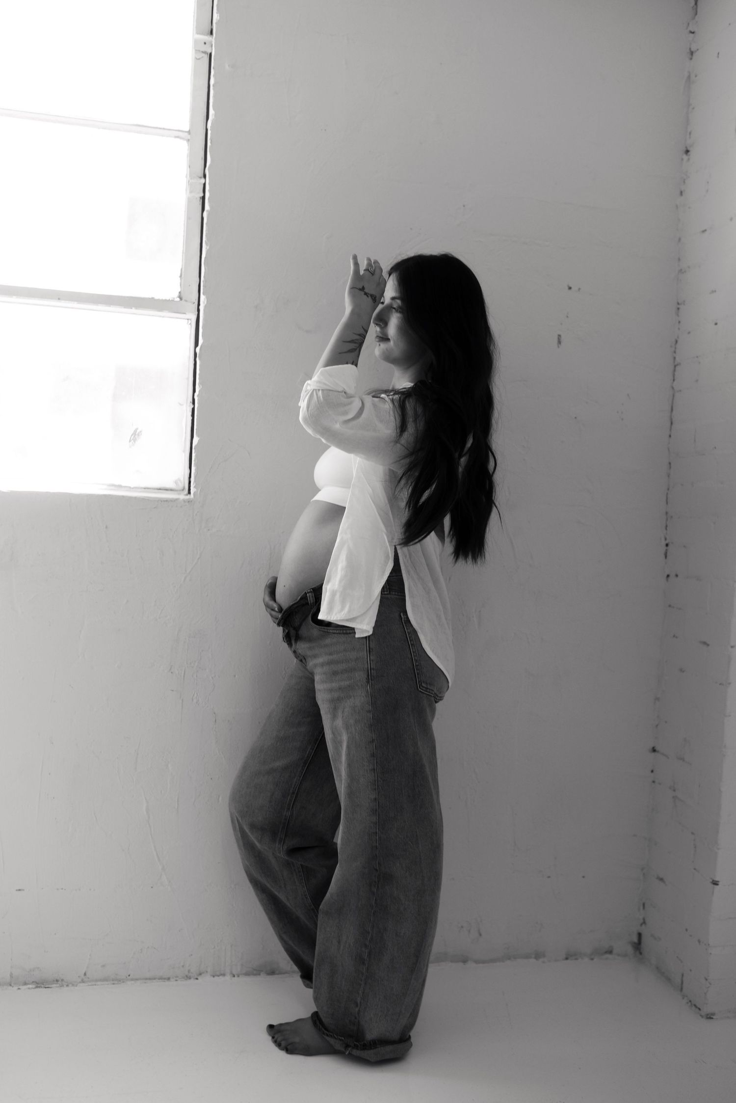 Black and white photo of a pregnant woman in white top and wide-leg pants leaning against a wall near a window.