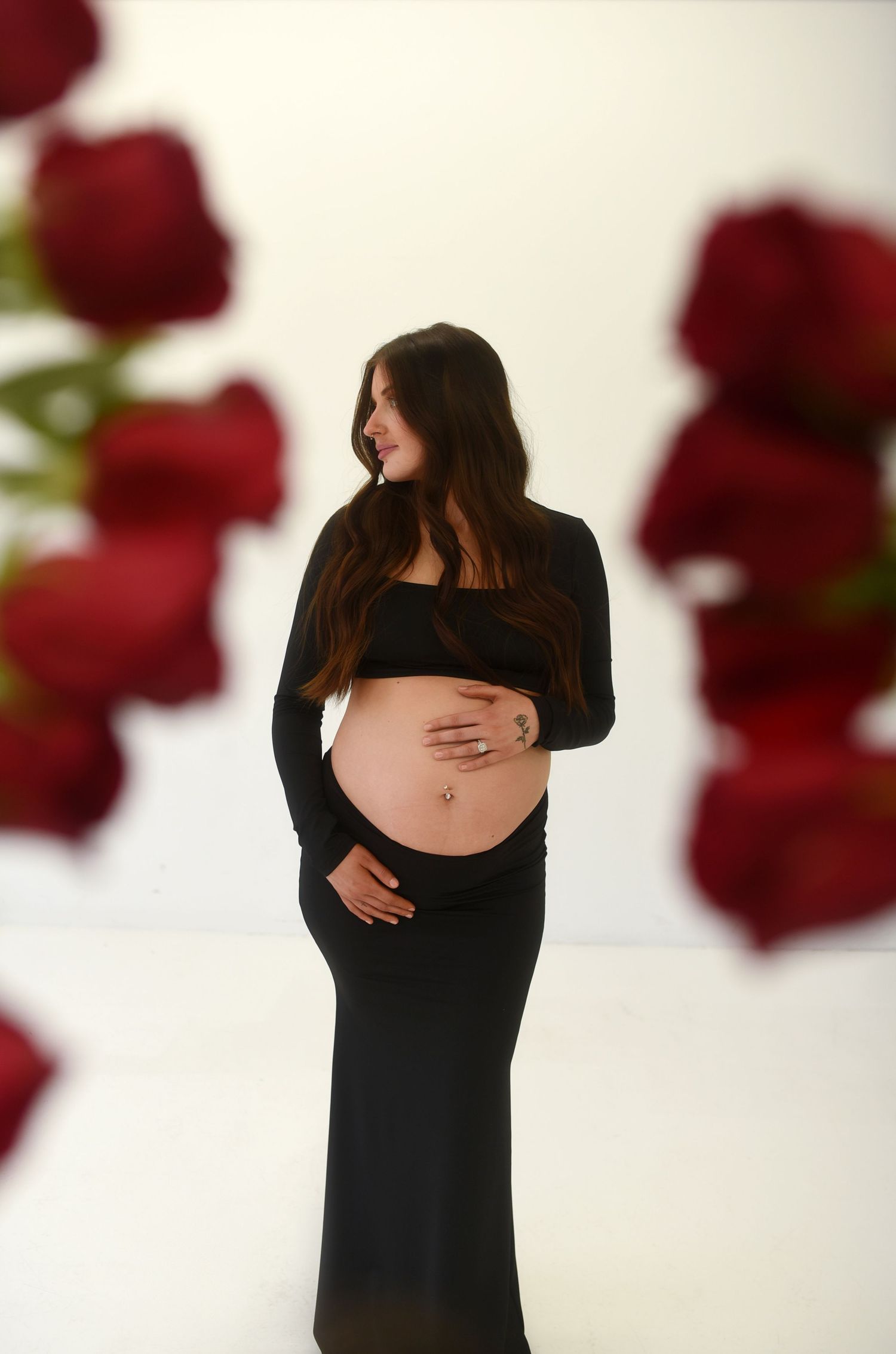 Pregnant woman in black crop top and skirt cradling baby bump, surrounded by blurred red roses on white background.