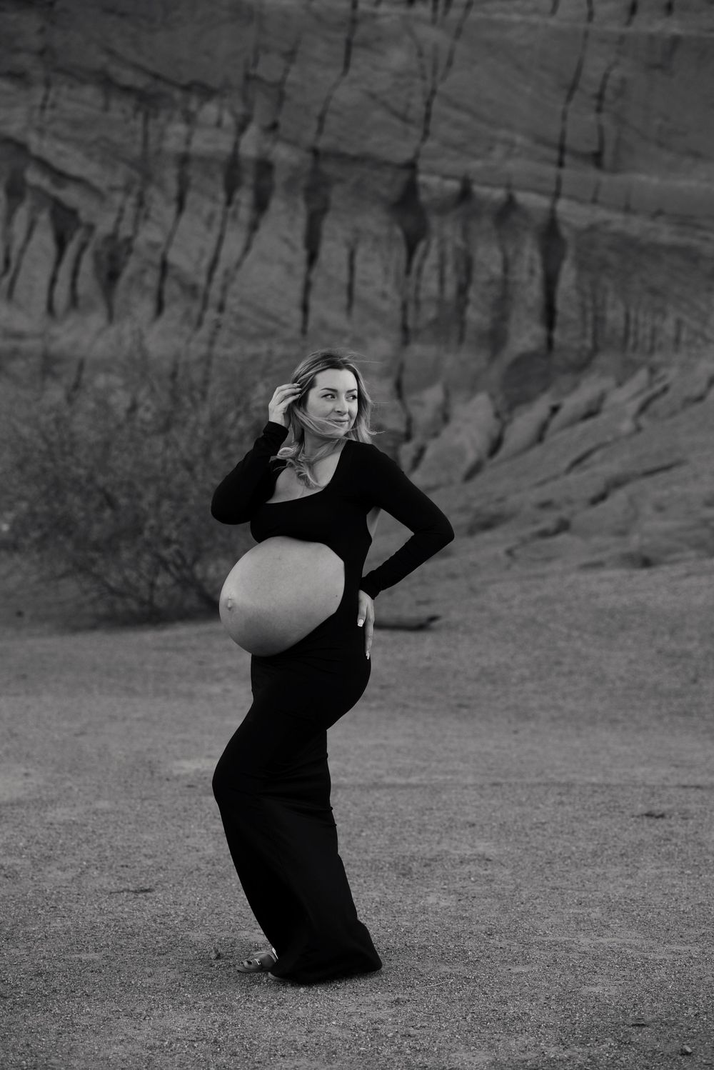 Black and white maternity photo of pregnant woman in black outfit posing in desert canyon landscape.