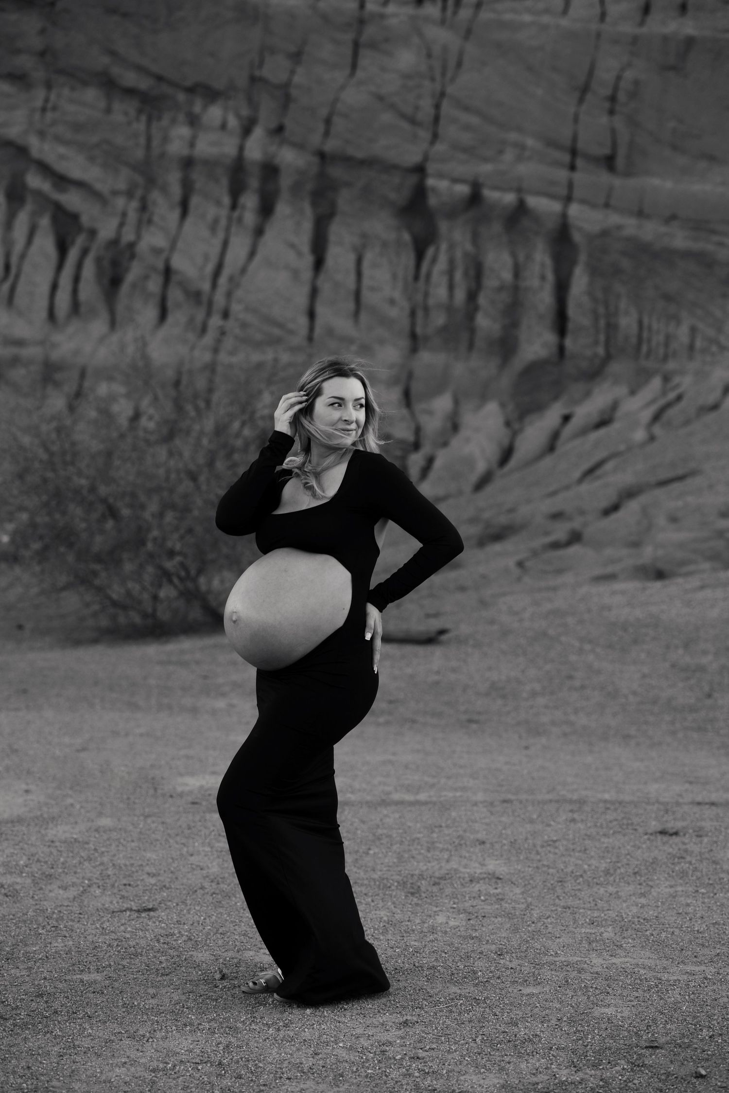 Black and white maternity photo of pregnant woman in black outfit posing in desert canyon landscape.
