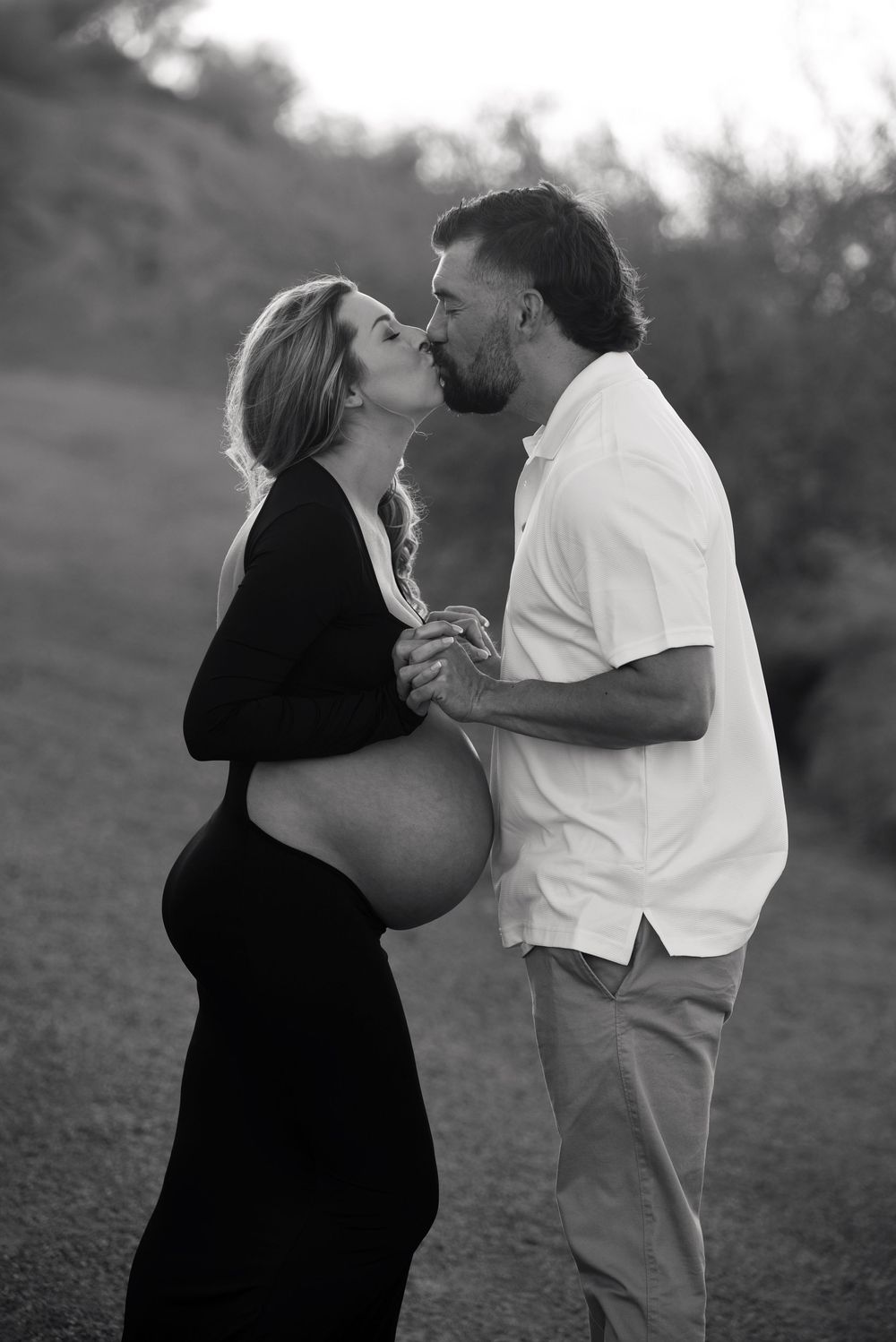 Black and white maternity photo of a couple kissing outdoors, woman in black dress showing baby bump.