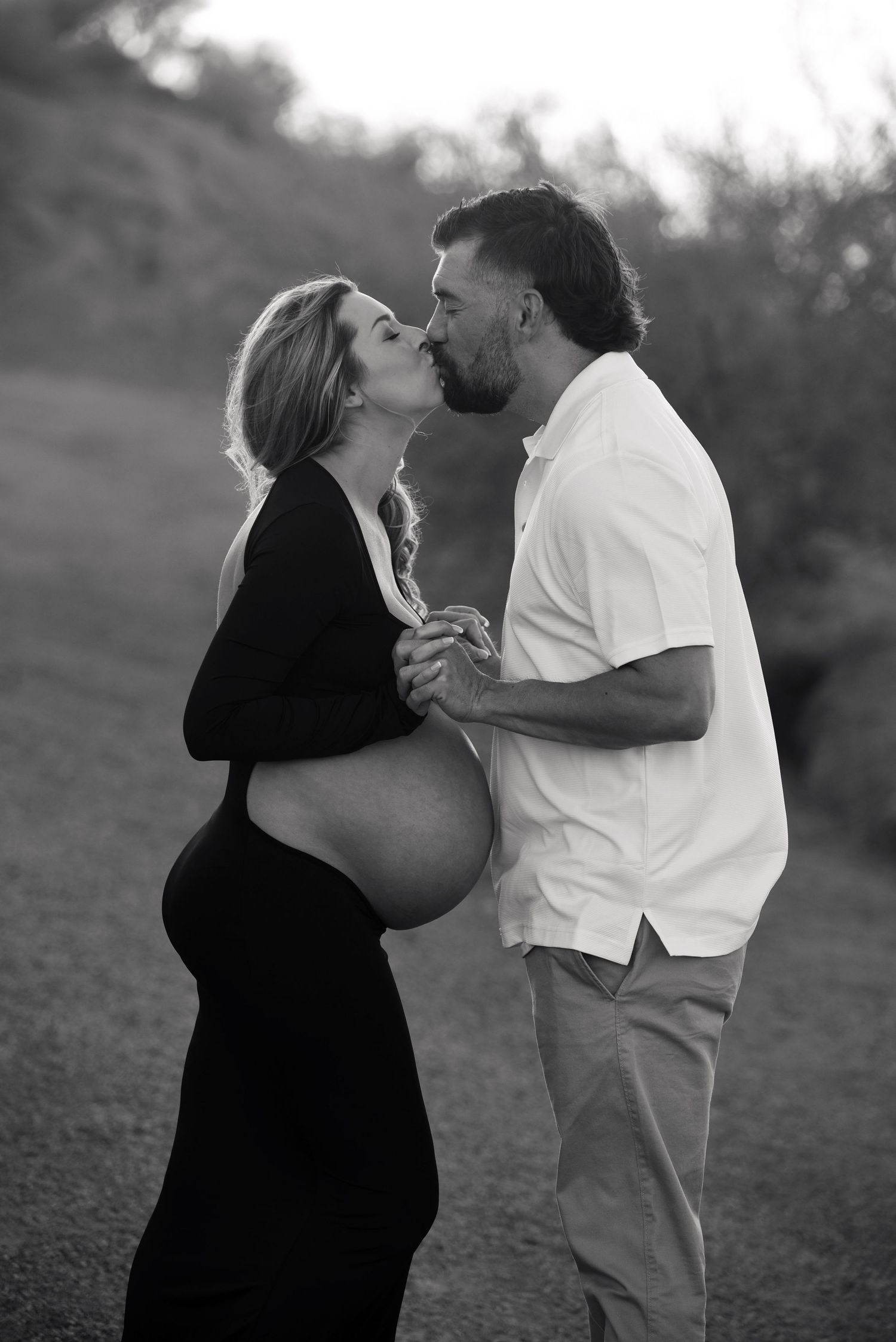Black and white maternity photo of a couple kissing outdoors, woman in black dress showing baby bump.