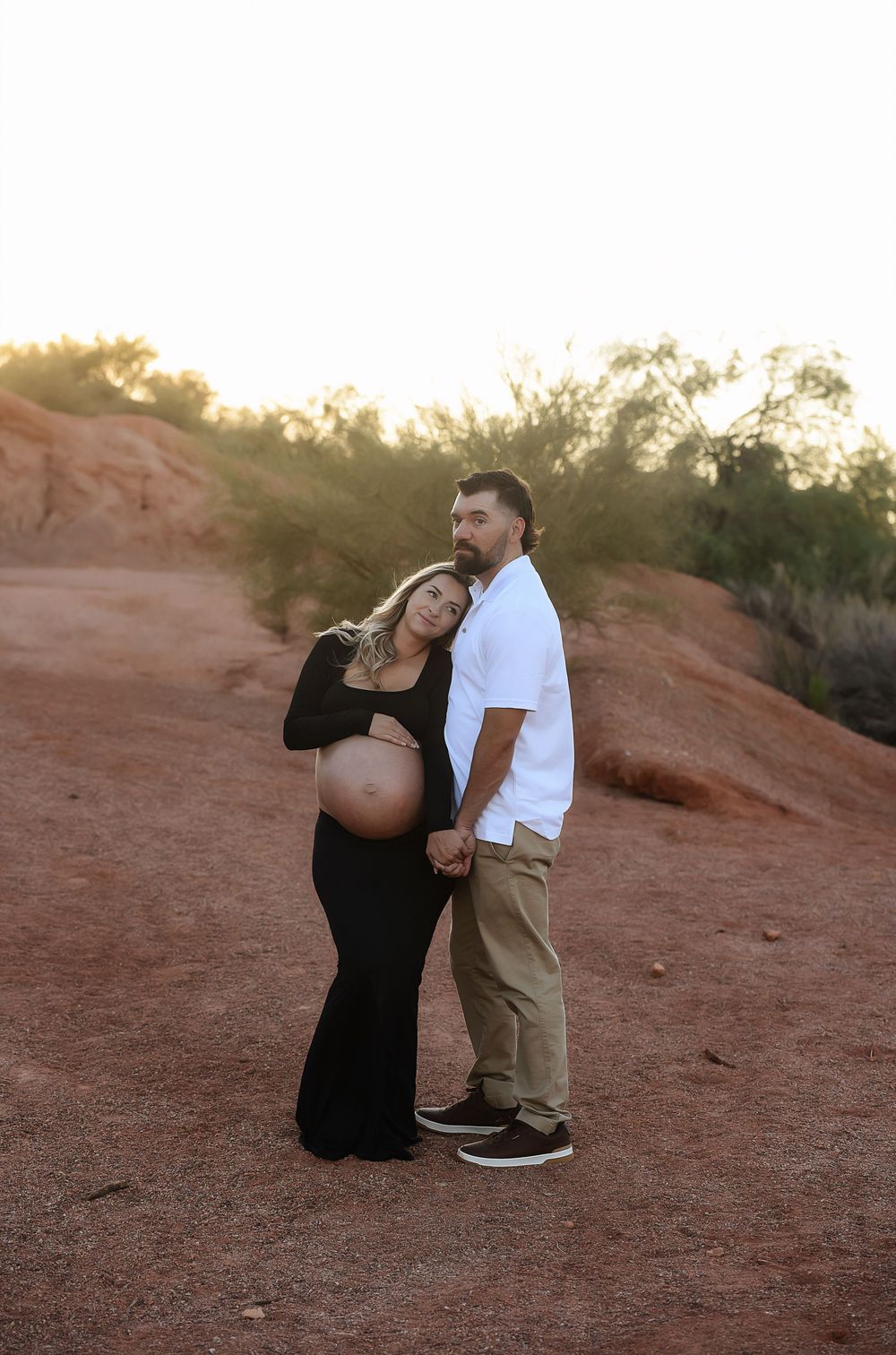 Pregnant couple poses at golden hour in desert landscape, woman in black maternity gown leaning on man in white shirt.