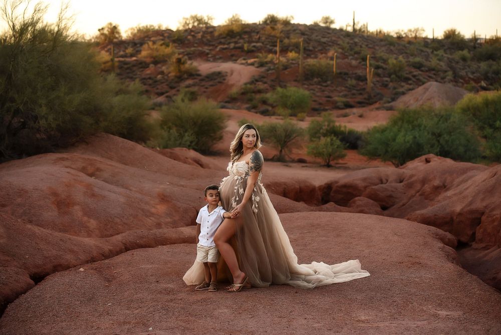 Pregnant woman in tulle gown poses with child on red rocks at sunset in Arizona desert landscape.