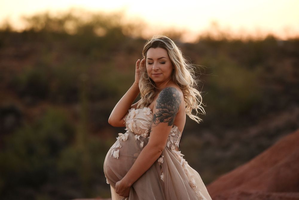 Pregnant woman in floral blush gown with tattoo poses outdoors at golden hour in desert landscape.
