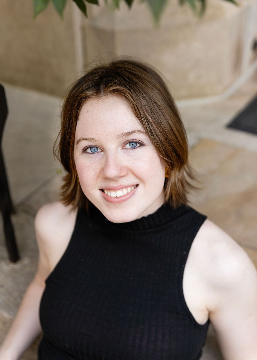Portrait of person in black sleeveless top with short brown hair against beige wall with plants.