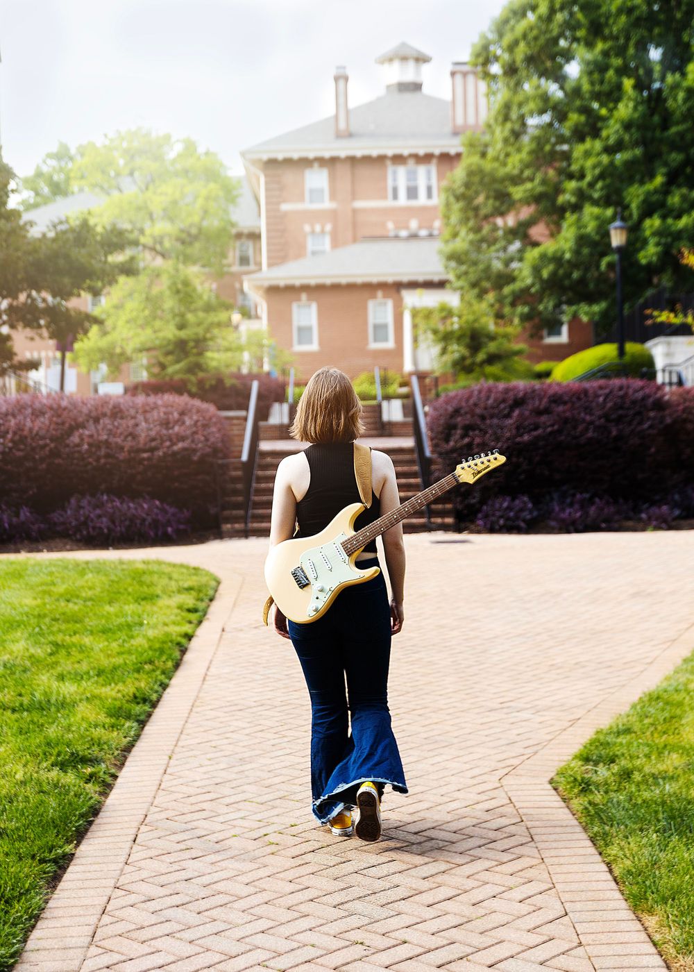 Person carrying electric guitar walking on campus pathway toward brick building.