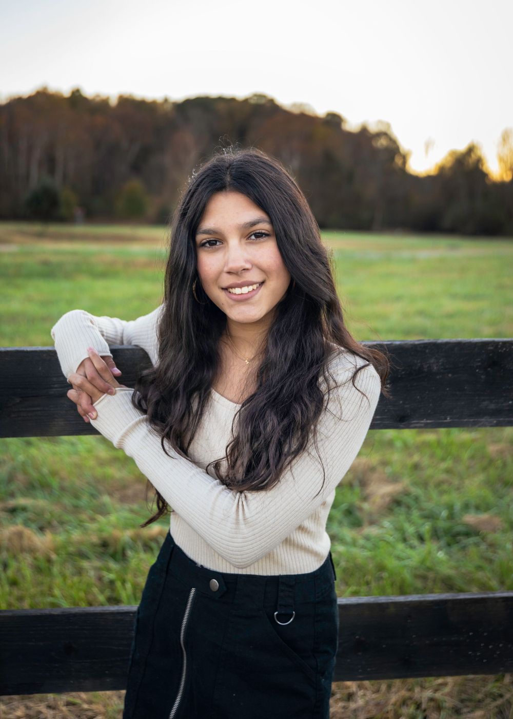 Portrait in white sweater leaning on wooden fence at sunset.