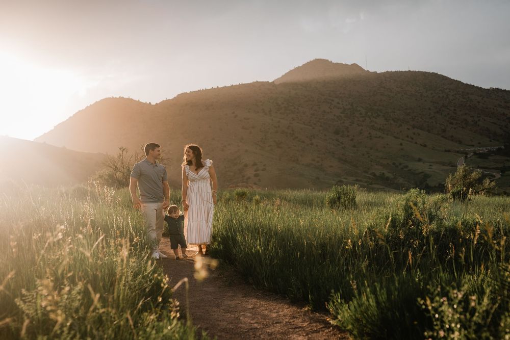 Family walking on a sunlit mountain trail surrounded by lush green fields at golden hour.