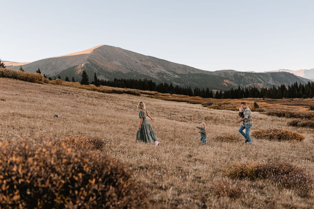 Family of three playing in a golden mountain meadow under clear skies with pine trees and peaks behind.