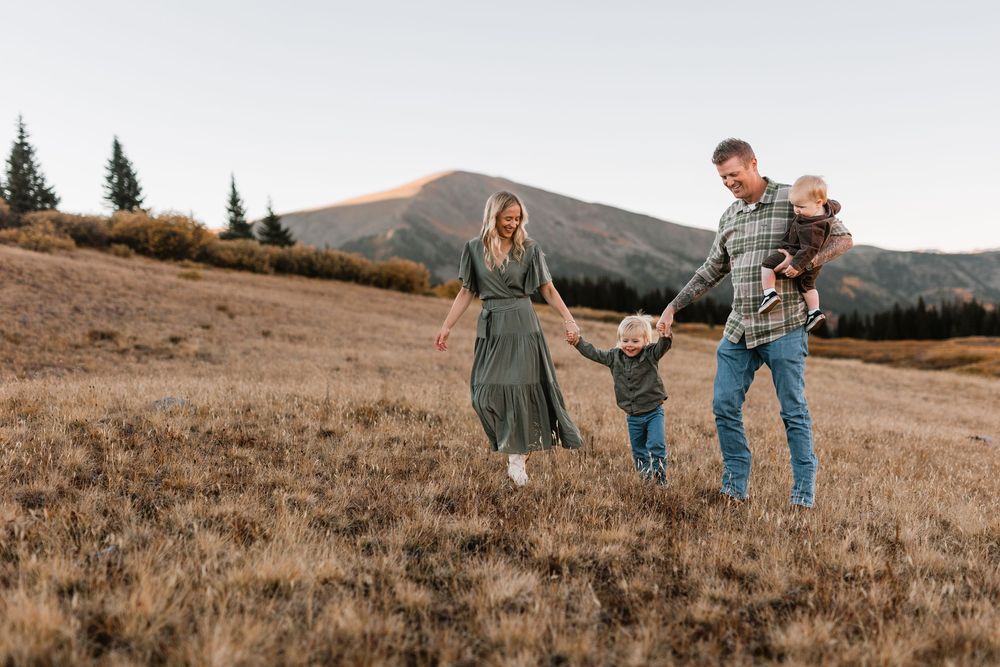 Family of four walking through golden mountain meadow, parents holding toddler's hands while dad carries baby.