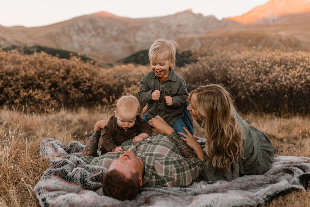 Family enjoying a golden hour picnic on a mountain meadow blanket with two young children playing.