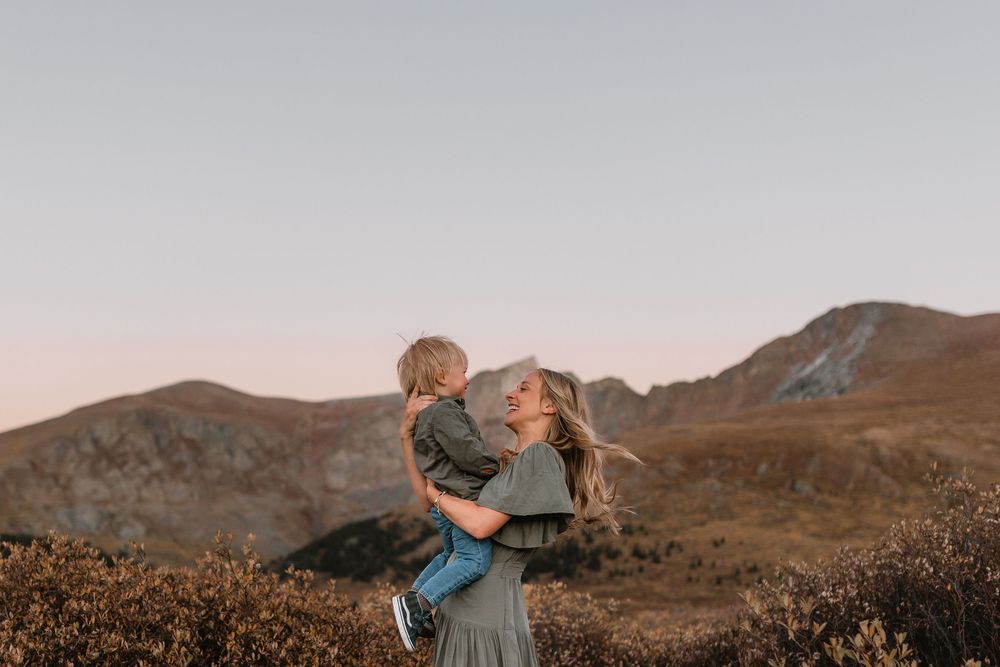 Mother lifting smiling toddler against scenic mountain backdrop at golden hour sunset.