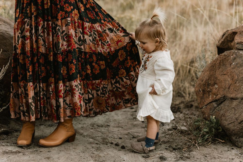Small child in white outfit standing next to floral dress and brown boots against rocks.