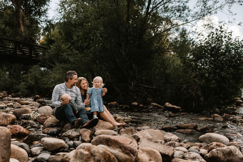 Family of four sitting on rocky riverbank surrounded by lush trees, sharing a joyful moment together outdoors.
