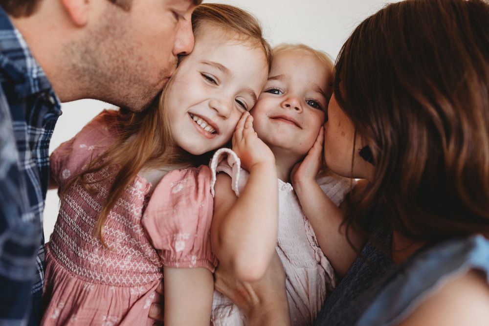 Happy family moment with parents kissing their two smiling young daughters on the cheeks indoors.