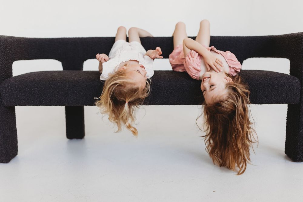 Two young girls hanging upside down over a dark bench, smiling and playing together against a white background.