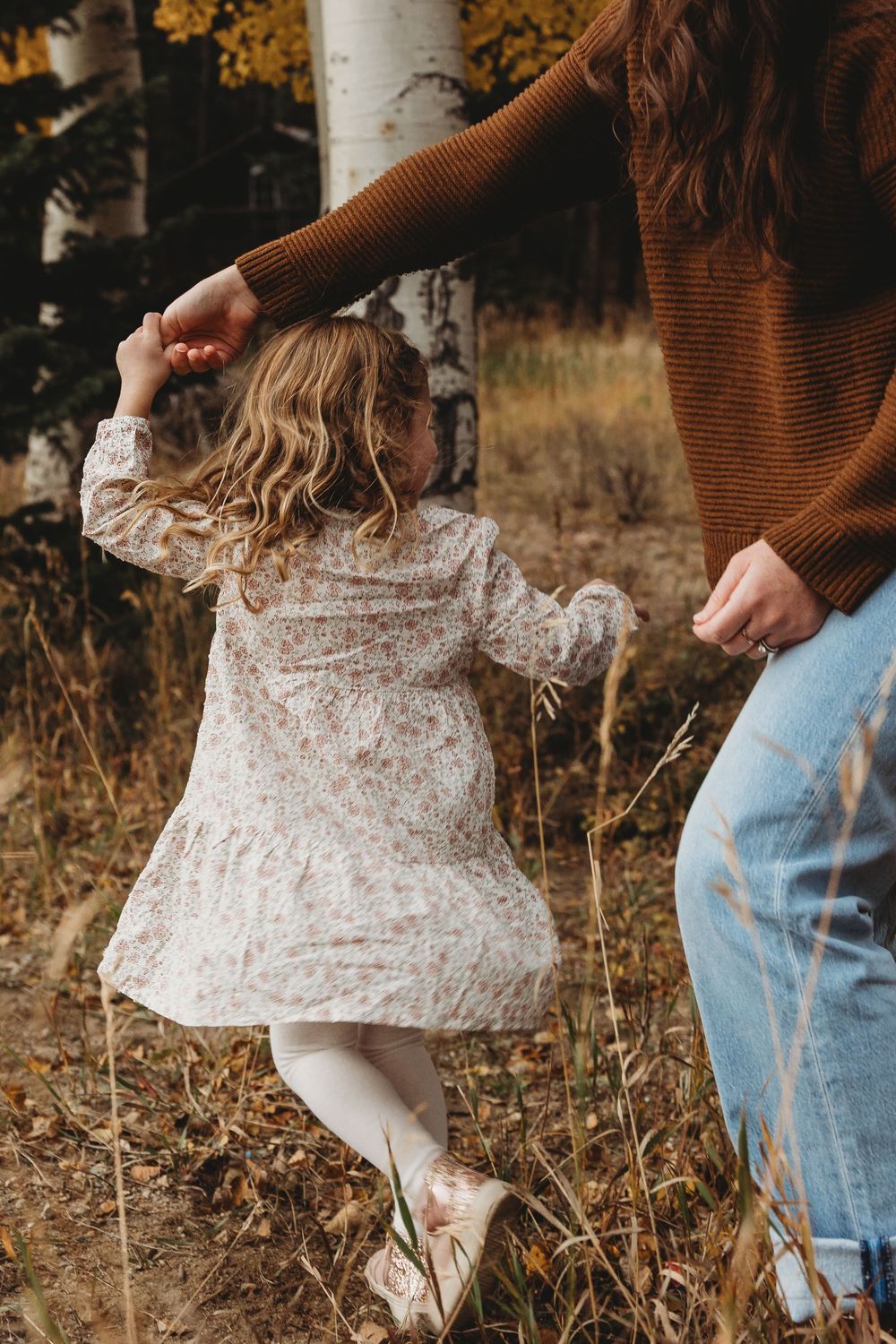Mother and daughter holding hands in autumn forest, girl in floral dress twirling among golden trees.