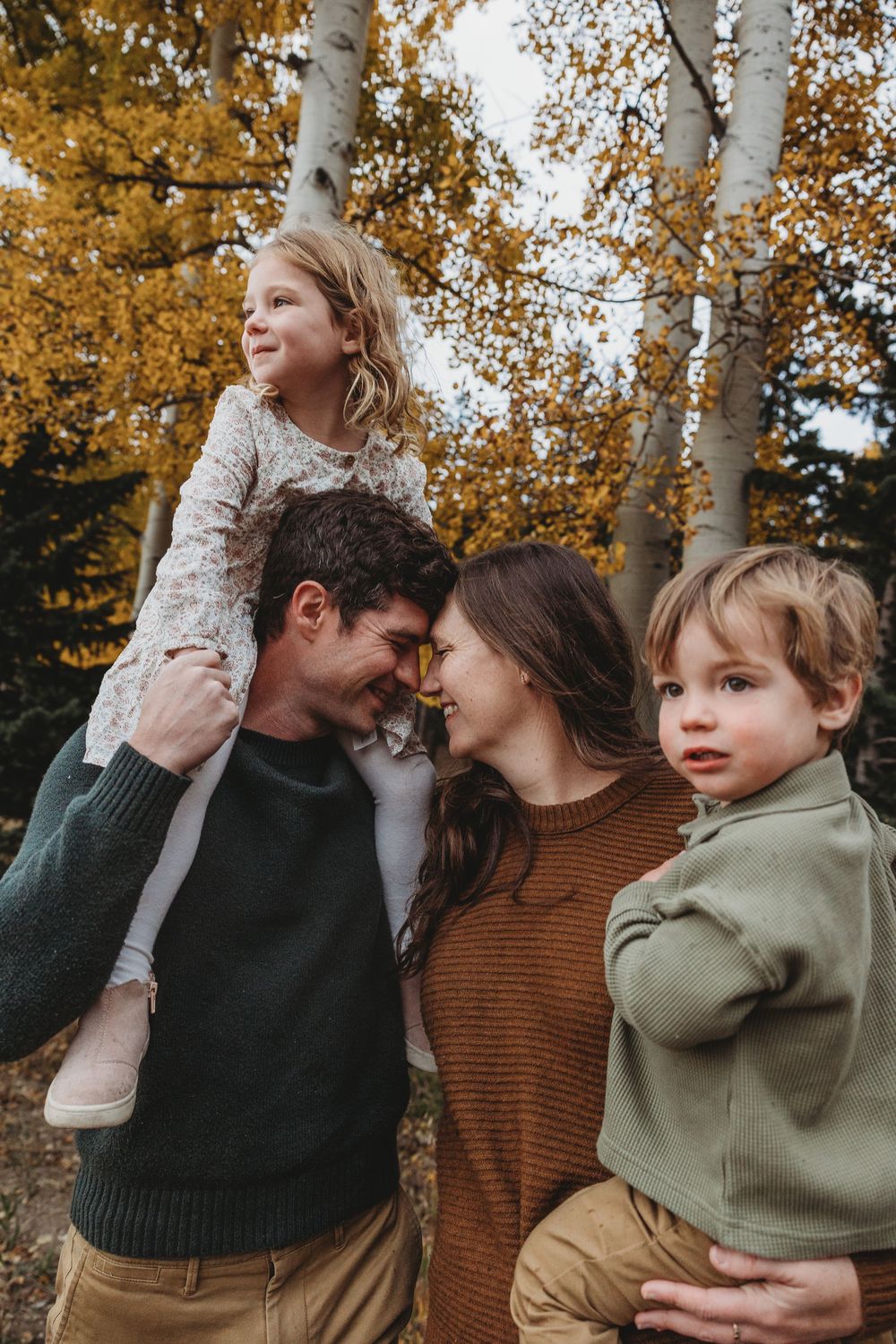 Family of four enjoying autumn outdoors, parents nuzzling noses while children sit on shoulders and hip.