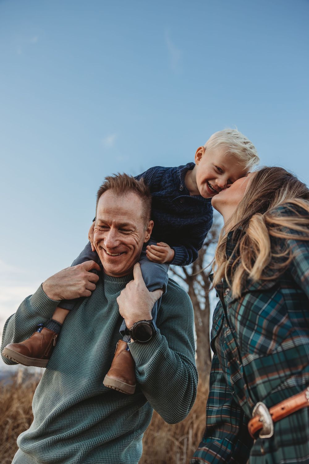 A happy family with a young blonde child sitting on dad's shoulders while mom smiles outdoors under a blue sky.