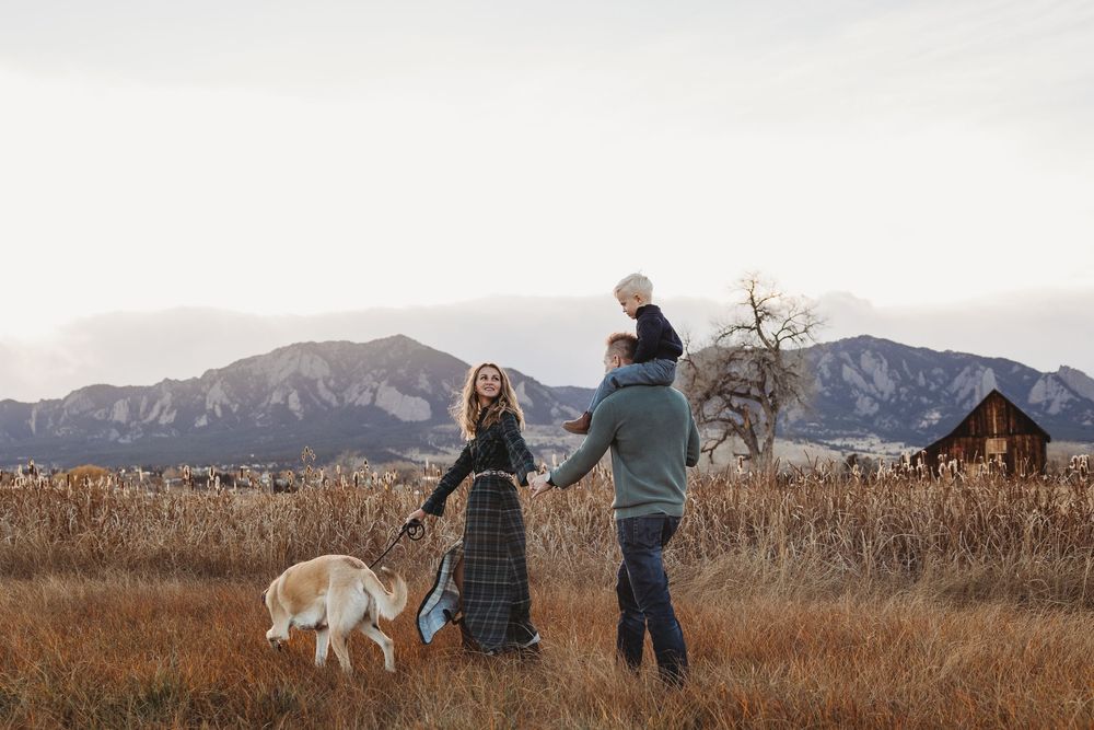 Family with dog walking in autumn field with mountains and rustic barn in background at sunset.