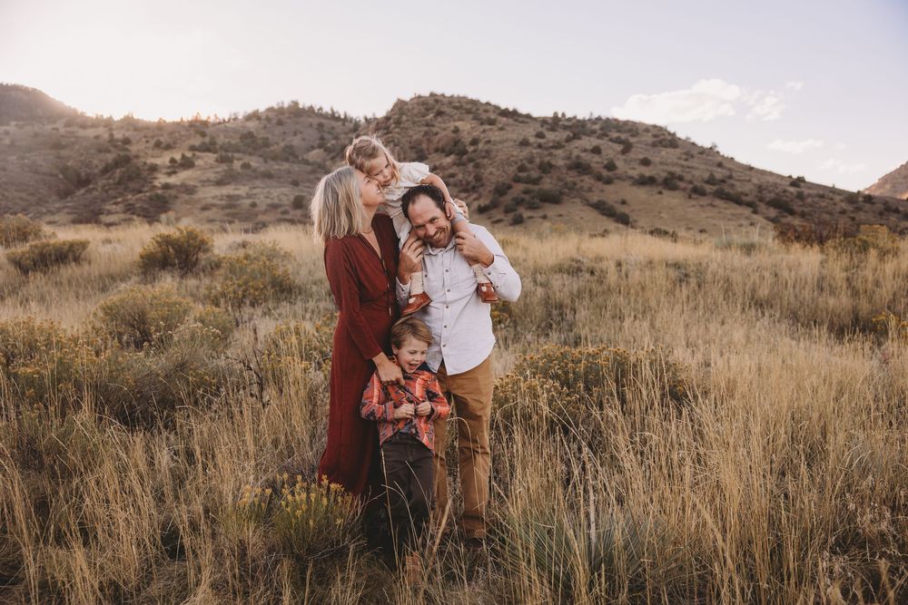 Family of four embracing in golden meadow with mountain backdrop during sunset photography session.