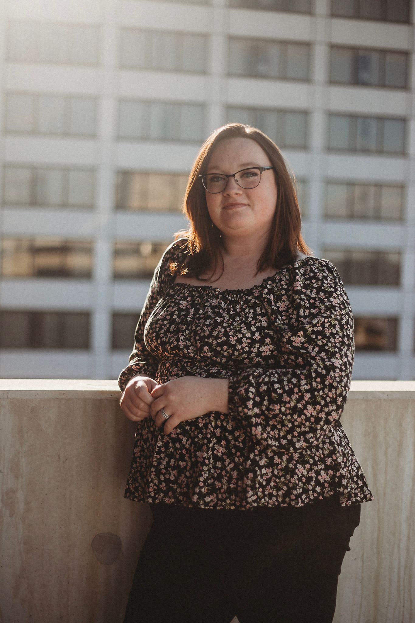 Professional portrait of someone in a floral print top posing against an urban building background at sunset.